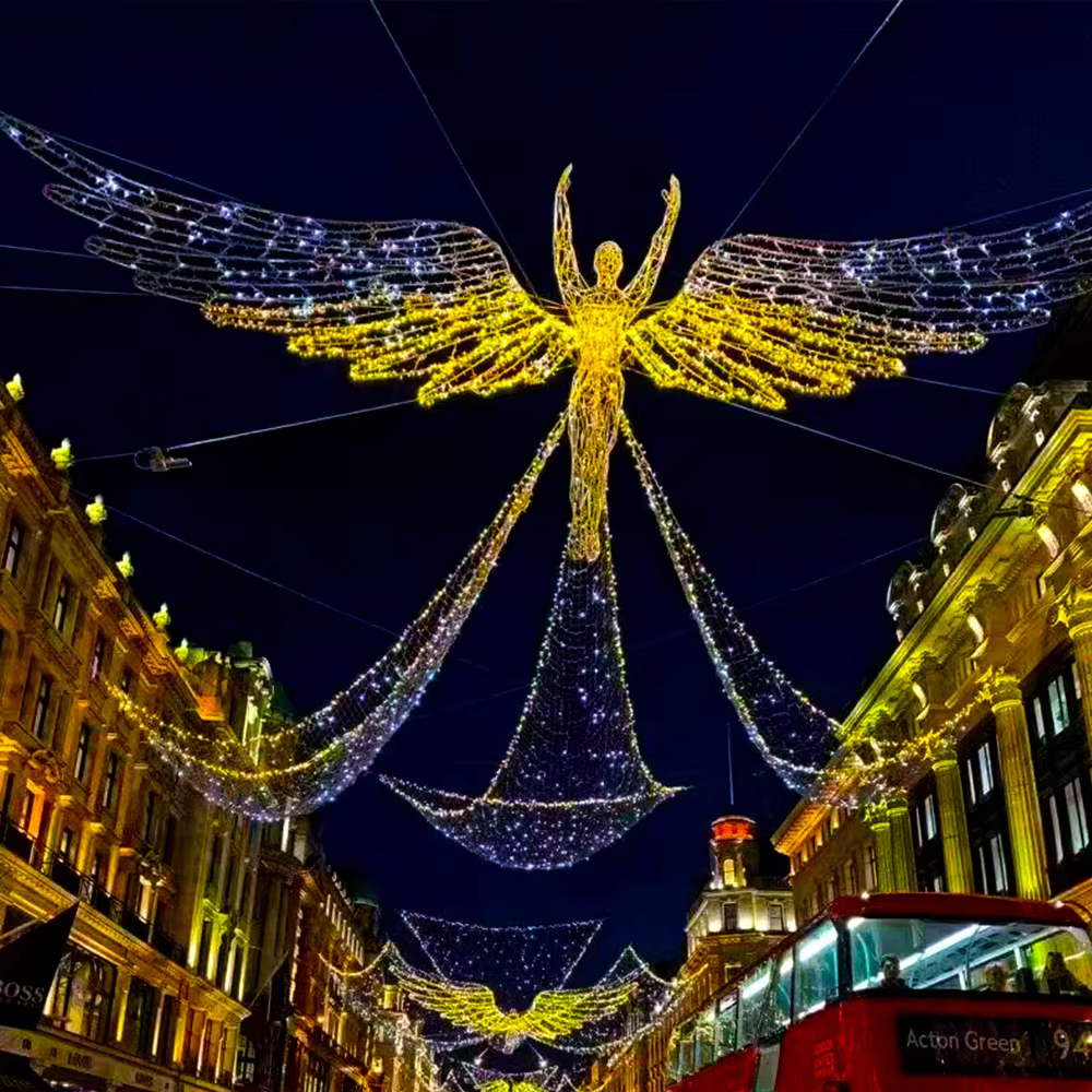L’image montre une grande installation lumineuse immersive en forme d’ange, suspendue au-dessus d’une rue, créant un spectaculaire décor lumineux immersif événementiel. Composé de milliers de petites lumières dorées et blanches, l’ange déploie ses ailes et semble flotter dans les airs, apportant une dimension magique à l’ensemble.Sa silhouette élancée et lumineuse diffuse une lumière chaleureuse qui enveloppe l’espace et crée une ambiance féérique ainsi qu’une atmosphère immersive. Répétées le long de la rue, ces décorations renforcent l’effet visuel et transforment l’environnement urbain en une véritable expérience sensorielle.Ce type de décor événementiel lumineux attire naturellement les regards et invite les visiteurs à s’arrêter, à contempler et à capturer des moments uniques. Il contribue à offrir une expérience événementielle mémorable, idéale pour dynamiser un lieu d’accueil ou un espace public.