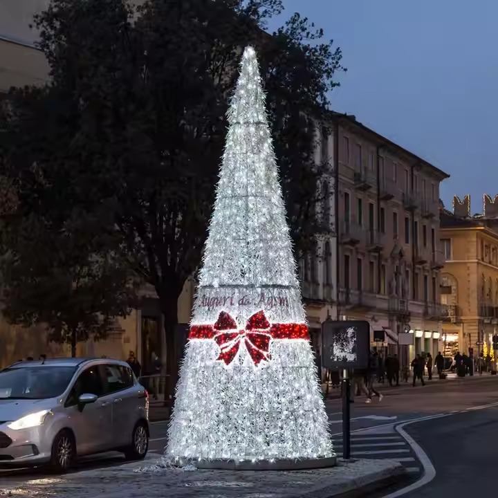 L’image présente un grand sapin de Noël lumineux installé au cœur d’un espace urbain, formant un décor lumineux immersif événementiel spectaculaire. Composé de milliers de petites lumières blanches scintillantes, il diffuse une lumière éclatante qui crée immédiatement une ambiance festive et immersive à la tombée du jour.Sa forme élancée et conique attire le regard et s’impose comme un point central dans la rue. Une large bande décorative entoure sa base, accompagnée d’un ruban lumineux rouge qui ajoute une touche chaleureuse et élégante à l’ensemble. Ce contraste visuel renforce l’atmosphère conviviale et met en valeur l’installation.Intégré dans un environnement urbain animé, entouré de bâtiments et de passants, ce décor événementiel lumineux transforme l’espace en une véritable expérience visuelle. Il invite les visiteurs à s’arrêter, à contempler et à capturer des moments uniques, contribuant ainsi à une expérience événementielle mémorable.Parfait pour dynamiser un centre-ville ou un lieu public, ce type d’installation lumineuse immersive crée une ambiance rassembleuse et festive, idéale pour la période des fêtes.