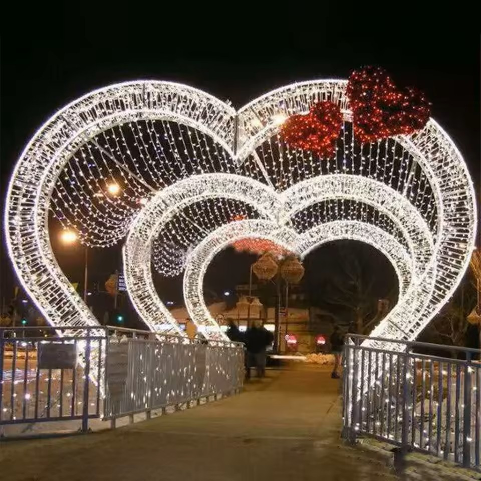 L’image met en scène un élégant tunnel lumineux en forme de cœurs, créant un spectaculaire décor lumineux immersif événementiel dans un environnement urbain. Composée de structures blanches illuminées, cette installation lumineuse immersive forme un passage enchanteur qui invite les visiteurs à traverser un univers empreint de douceur et de romantisme.Les arches en forme de cœurs, rehaussées de délicates guirlandes lumineuses et d’un accent rouge décoratif, diffusent une lumière chaleureuse qui enveloppe l’espace et instaure une ambiance intime et féérique. La perspective répétée des structures accentue l’atmosphère immersive, transformant ce simple pont en une véritable expérience visuelle.Ce type de décor événementiel lumineux attire naturellement les passants, favorise les arrêts spontanés et devient un lieu privilégié pour la prise de photos. Il contribue à créer une expérience événementielle mémorable, idéale pour les espaces publics, les lieux d’accueil ou les événements saisonniers.