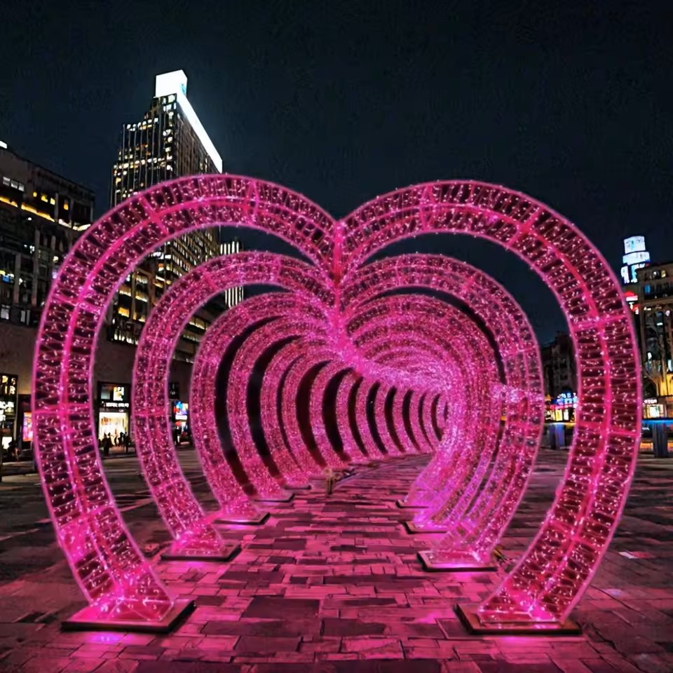 L’image présente un tunnel lumineux en forme de cœurs installé en plein espace urbain, créant un spectaculaire décor lumineux immersif événementiel. Composée de plusieurs arches roses illuminées, cette installation lumineuse immersive forme un passage qui invite les visiteurs à circuler au cœur d’une ambiance romantique et chaleureuse.La répétition des structures crée une perspective profonde et enveloppante, renforçant l’atmosphère immersive et l’effet visuel. La lumière rosée se reflète sur le sol pavé, accentuant le caractère intime et poétique du lieu. Ce type de décor événementiel lumineux transforme l’espace en une véritable expérience sensorielle, propice à la contemplation, aux échanges et à la prise de photos.Parfait pour un lieu d’accueil ou un espace public, ce décor lumineux immersif attire naturellement les regards et favorise l’interaction. Il offre une expérience événementielle mémorable, idéale pour créer un moment unique et marquer les esprits.